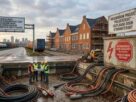 Construction workers in high-visibility vests study technical plans inside a concrete utility trench filled with thick, disconnected power cables, set against a newly built Dutch residential development and grid capacity warning signage.