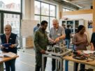 A group of adult newcomers in a Dutch vocational training workshop engaging in hands-on learning with instructors, set against large windows overlooking canal houses.